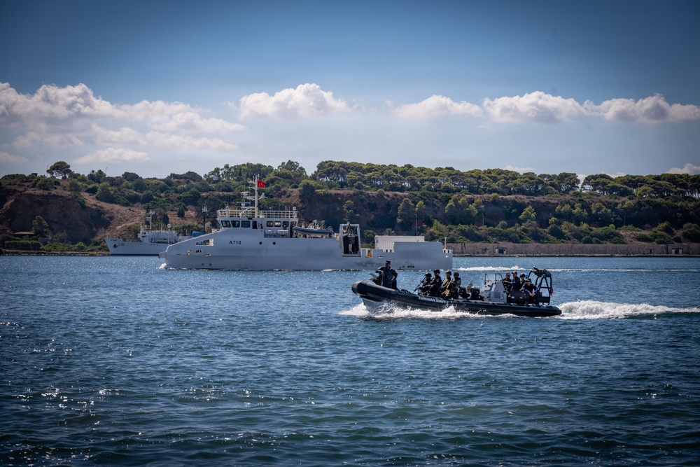 A military or naval operation is underway on open water, featuring a rigid inflatable boat (RIB) carrying armed personnel in the foreground and a larger naval vessel marked 'A710' in the background. A red flag, consistent with the Turkish national flag, is visible on the larger ship, suggesting Turkish naval affiliation. The scene appears to be a maritime exercise or patrol operation near a coastline with dense tree coverage.