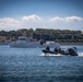 A military or naval operation is underway on open water, featuring a rigid inflatable boat (RIB) carrying armed personnel in the foreground and a larger naval vessel marked 'A710' in the background. A red flag, consistent with the Turkish national flag, is visible on the larger ship, suggesting Turkish naval affiliation. The scene appears to be a maritime exercise or patrol operation near a coastline with dense tree coverage.