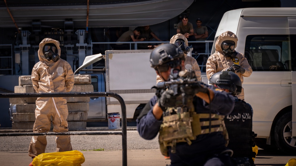 The image depicts what appears to be a military or law enforcement training exercise involving CBRN (Chemical, Biological, Radiological, Nuclear) response personnel in tan hazmat suits with gas masks, alongside armed tactical operators in body armor and helmets. The scene takes place in an outdoor area near vehicles and a dock or port facility. Arabic text visible on one operator's vest reads 'طلائع البحرية' (Naval Vanguards), suggesting a Middle Eastern naval or coast guard unit.
