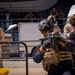 The image depicts what appears to be a military or law enforcement training exercise involving CBRN (Chemical, Biological, Radiological, Nuclear) response personnel in tan hazmat suits with gas masks, alongside armed tactical operators in body armor and helmets. The scene takes place in an outdoor area near vehicles and a dock or port facility. Arabic text visible on one operator's vest reads 'طلائع البحرية' (Naval Vanguards), suggesting a Middle Eastern naval or coast guard unit.
