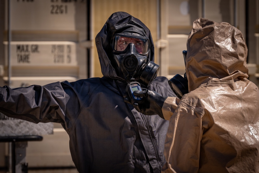 Two individuals wearing full hazmat/CBRN protective gear are engaged in what appears to be a decontamination or radiation survey procedure. The person in a black protective suit and gas mask is holding a handheld detection device, scanning or checking the person in a tan/gold protective suit. The setting appears to be near shipping containers, suggesting a training exercise or emergency response scenario.