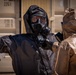 Two individuals wearing full hazmat/CBRN protective gear are engaged in what appears to be a decontamination or radiation survey procedure. The person in a black protective suit and gas mask is holding a handheld detection device, scanning or checking the person in a tan/gold protective suit. The setting appears to be near shipping containers, suggesting a training exercise or emergency response scenario.