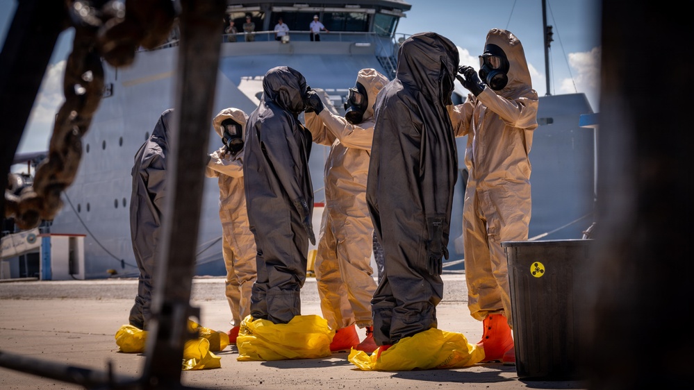 Personnel in full CBRN (Chemical, Biological, Radiological, and Nuclear) protective gear are conducting what appears to be a training exercise or decontamination drill at a port facility. The individuals are wearing hazmat suits in tan and dark gray colors, full-face gas masks, yellow boot covers, and orange overshoes. A large white vessel is docked in the background with observers watching from an upper deck.