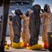 Personnel in full CBRN (Chemical, Biological, Radiological, and Nuclear) protective gear are conducting what appears to be a training exercise or decontamination drill at a port facility. The individuals are wearing hazmat suits in tan and dark gray colors, full-face gas masks, yellow boot covers, and orange overshoes. A large white vessel is docked in the background with observers watching from an upper deck.