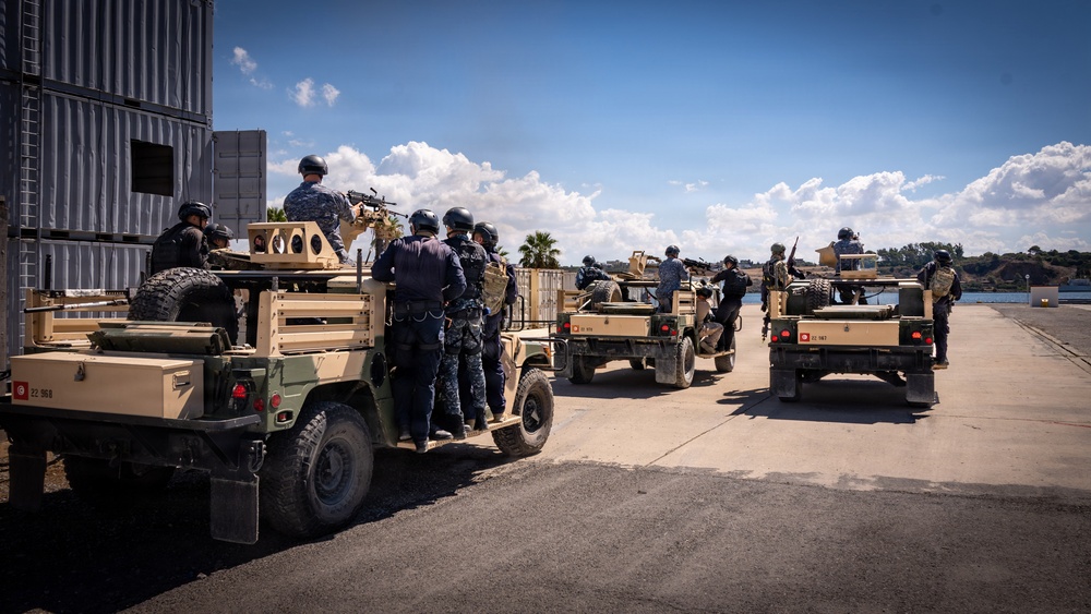 A group of military or security personnel in tactical gear and helmets are gathered around multiple tan/desert-colored Humvee-type military vehicles on a paved waterfront area. The vehicles appear to be equipped with weapon mounts on top, and personnel are positioned around and on the vehicles in what appears to be a training or operational exercise. The setting includes a body of water in the background, blue sky with clouds, and what appears to be a port or naval facility.