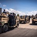 A group of military or security personnel in tactical gear and helmets are gathered around multiple tan/desert-colored Humvee-type military vehicles on a paved waterfront area. The vehicles appear to be equipped with weapon mounts on top, and personnel are positioned around and on the vehicles in what appears to be a training or operational exercise. The setting includes a body of water in the background, blue sky with clouds, and what appears to be a port or naval facility.