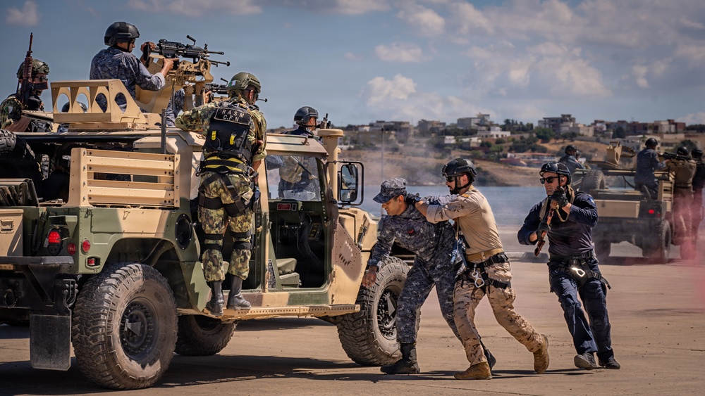 The image depicts a military tactical exercise or demonstration involving multiple personnel in varied uniforms operating around a tan/desert-colored Humvee (HMMWV). Personnel are engaged in what appears to be a coordinated scenario including a simulated detainee handling or casualty extraction near the vehicle. Pink smoke is visible in the background, suggesting a training or demonstration environment.