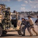 The image depicts a military tactical exercise or demonstration involving multiple personnel in varied uniforms operating around a tan/desert-colored Humvee (HMMWV). Personnel are engaged in what appears to be a coordinated scenario including a simulated detainee handling or casualty extraction near the vehicle. Pink smoke is visible in the background, suggesting a training or demonstration environment.