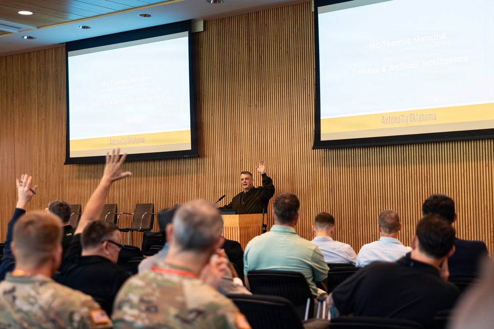 A presenter stands at a podium with one hand raised, addressing an audience in a modern conference room. Two projection screens display slides referencing 'MG Thomas Mansino' and 'Defense &amp; Artificial Intelligence' under the branding 'Autonomy Oklahoma'. Several audience members, including individuals in military uniforms, have raised their hands in response to the presenter.