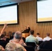 A presenter stands at a podium with one hand raised, addressing an audience in a modern conference room. Two projection screens display slides referencing 'MG Thomas Mansino' and 'Defense &amp; Artificial Intelligence' under the branding 'Autonomy Oklahoma'. Several audience members, including individuals in military uniforms, have raised their hands in response to the presenter.