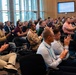 A large audience is seated in a modern conference or auditorium room, with the majority of attendees visibly applauding. The crowd is diverse and includes individuals in business attire, military uniforms, and casual clothing, many wearing conference lanyards and badges. A presentation screen is visible at the back of the room displaying text content.