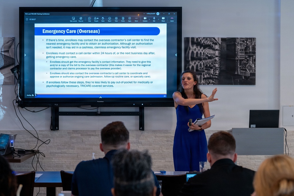 A woman in a blue dress is presenting at a POC and TRICARE Training Conference, standing beside a large display screen showing a slide titled 'Emergency Care (Overseas)'. The presentation appears to be conducted via a video conferencing platform (likely Microsoft Teams) and covers TRICARE overseas emergency care procedures. Several attendees are seated in the foreground with their backs to the camera.