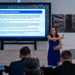 A woman in a blue dress is presenting at a POC and TRICARE Training Conference, standing beside a large display screen showing a slide titled 'Emergency Care (Overseas)'. The presentation appears to be conducted via a video conferencing platform (likely Microsoft Teams) and covers TRICARE overseas emergency care procedures. Several attendees are seated in the foreground with their backs to the camera.
