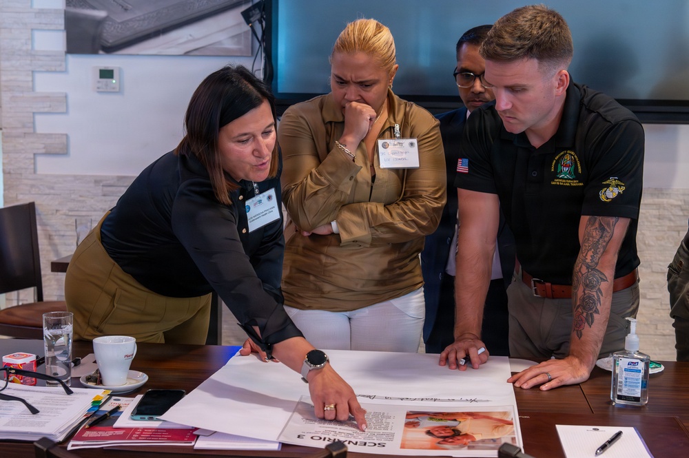 Four individuals are gathered around a conference table, intently reviewing a document labeled 'SCENARIO 3' that includes printed text and a photograph. The group appears to be engaged in a training exercise or workshop, with one woman pointing to specific details on the document. The setting appears to be a professional meeting or briefing room.