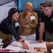 Four individuals are gathered around a conference table, intently reviewing a document labeled 'SCENARIO 3' that includes printed text and a photograph. The group appears to be engaged in a training exercise or workshop, with one woman pointing to specific details on the document. The setting appears to be a professional meeting or briefing room.