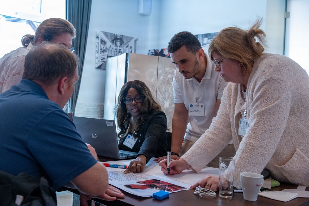A group of five people are gathered around a table in what appears to be a professional workshop or collaborative meeting setting. They are actively engaged in reviewing and annotating documents spread across the table, with a Dell laptop open among them. Several participants are wearing name badges, suggesting a formal conference or organized event.
