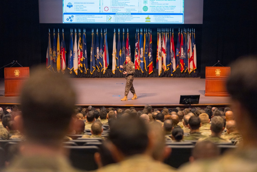 A military officer in camouflage uniform is presenting on stage to a large audience of uniformed military personnel in an auditorium. The stage is flanked by numerous military and U.S. state flags, with two wooden podiums bearing an emblem on either side. A presentation slide titled 'Administration' is projected on a screen behind the speaker.