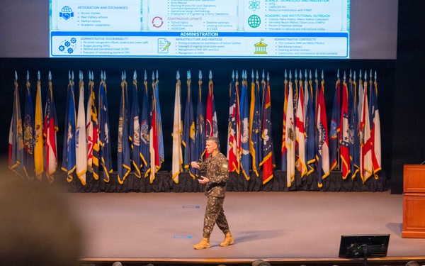 A military officer in camouflage uniform is presenting on stage to a large audience of uniformed military personnel in an auditorium. The stage is flanked by numerous military and U.S. state flags, with two wooden podiums bearing an emblem on either side. A presentation slide titled 'Administration' is projected on a screen behind the speaker.