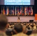 A military officer in camouflage uniform is presenting on stage to a large audience of uniformed military personnel in an auditorium. The stage is flanked by numerous military and U.S. state flags, with two wooden podiums bearing an emblem on either side. A presentation slide titled 'Administration' is projected on a screen behind the speaker.