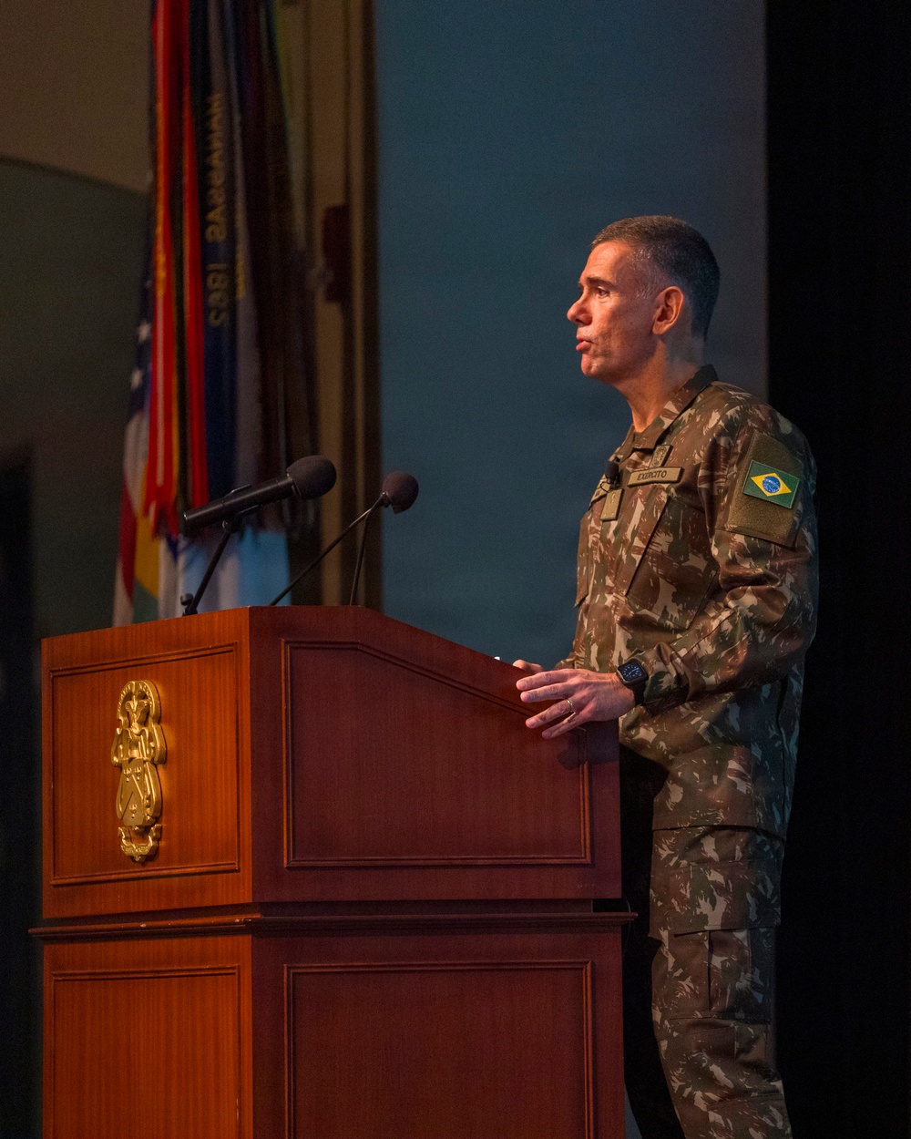 A Brazilian Army officer in camouflage uniform stands at a wooden podium delivering a speech or presentation. The podium bears a gold emblem, and multiple flags are visible in the background. The setting appears to be a formal military auditorium or conference hall.