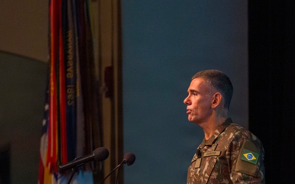 A Brazilian Army officer in camouflage uniform stands at a wooden podium delivering a speech or presentation. The podium bears a gold emblem, and multiple flags are visible in the background. The setting appears to be a formal military auditorium or conference hall.