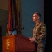 A Brazilian Army officer in camouflage uniform stands at a wooden podium delivering a speech or presentation. The podium bears a gold emblem, and multiple flags are visible in the background. The setting appears to be a formal military auditorium or conference hall.