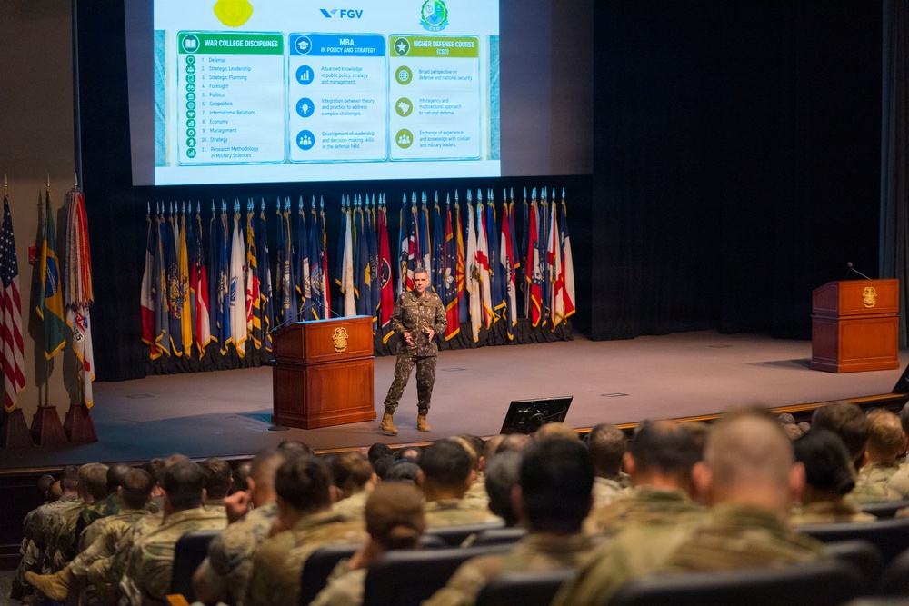 A military officer in camouflage uniform delivers a presentation at a podium on a formal auditorium stage, with a large projection screen displaying a slide about 'War College Disciplines,' 'MBA in Policy and Strategy,' and 'Higher Defense Course' under the FGV branding. The audience consists of numerous military personnel in uniform seated in rows. The backdrop features a large display of military and state flags.