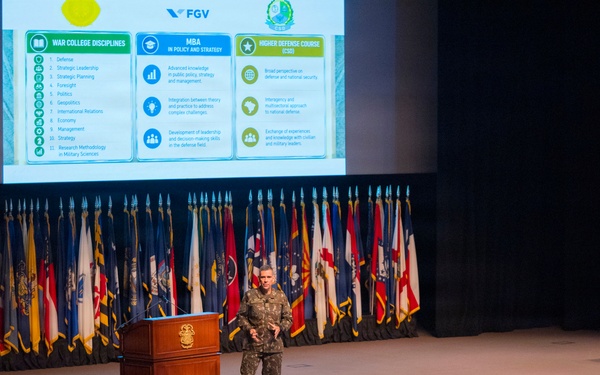 A military officer in camouflage uniform delivers a presentation at a podium on a formal auditorium stage, with a large projection screen displaying a slide about 'War College Disciplines,' 'MBA in Policy and Strategy,' and 'Higher Defense Course' under the FGV branding. The audience consists of numerous military personnel in uniform seated in rows. The backdrop features a large display of military and state flags.