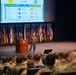 A military officer in camouflage uniform delivers a presentation at a podium on a formal auditorium stage, with a large projection screen displaying a slide about 'War College Disciplines,' 'MBA in Policy and Strategy,' and 'Higher Defense Course' under the FGV branding. The audience consists of numerous military personnel in uniform seated in rows. The backdrop features a large display of military and state flags.