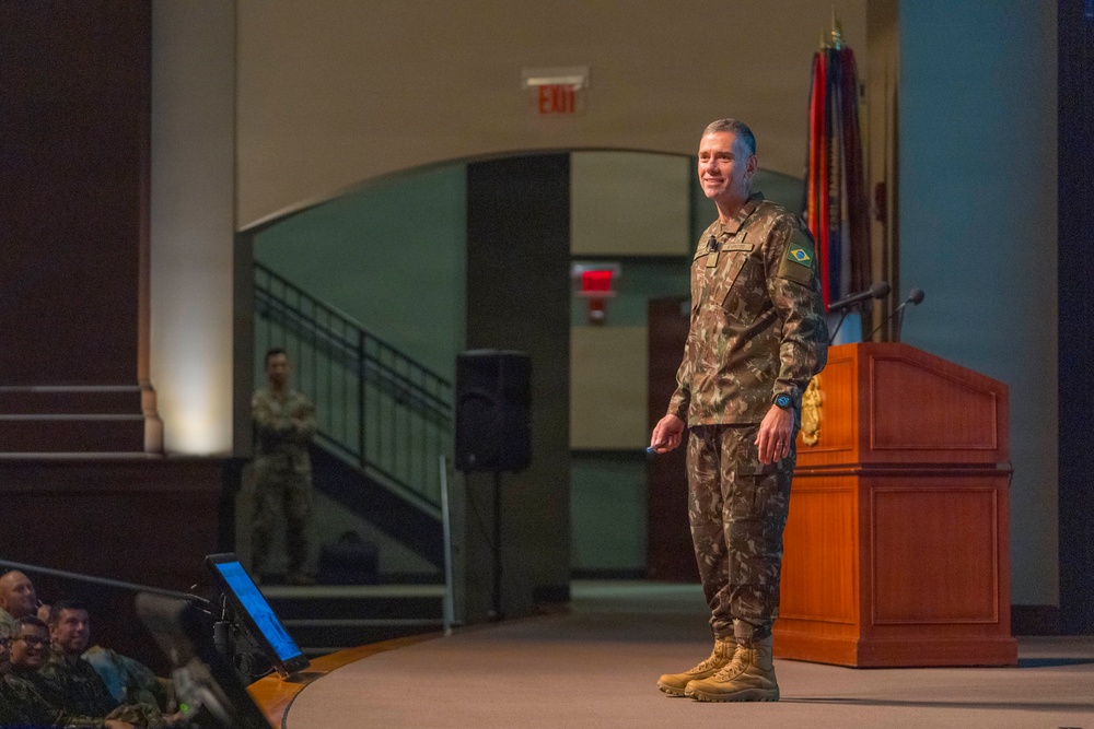 A military officer wearing Brazilian Army camouflage uniform stands on a stage near a wooden podium, apparently delivering a presentation or lecture to an audience. The setting appears to be a formal military auditorium or conference hall, with flags visible in the background. A second uniformed soldier stands in the background near a staircase.