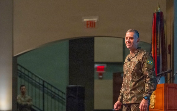 A military officer wearing Brazilian Army camouflage uniform stands on a stage near a wooden podium, apparently delivering a presentation or lecture to an audience. The setting appears to be a formal military auditorium or conference hall, with flags visible in the background. A second uniformed soldier stands in the background near a staircase.