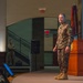 A military officer wearing Brazilian Army camouflage uniform stands on a stage near a wooden podium, apparently delivering a presentation or lecture to an audience. The setting appears to be a formal military auditorium or conference hall, with flags visible in the background. A second uniformed soldier stands in the background near a staircase.
