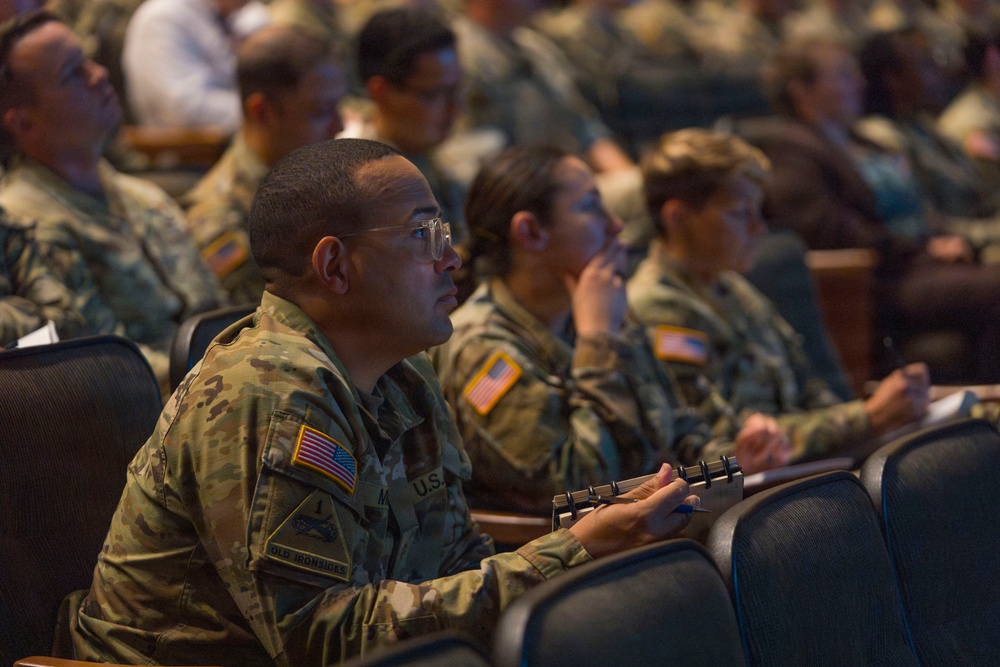 A large group of U.S. Army soldiers in OCP (Operational Camouflage Pattern) uniforms are seated in an auditorium-style venue, attentively watching a presentation or briefing. The foreground soldier is wearing glasses and holding a small notebook, suggesting active note-taking. The setting appears to be a formal military conference, training event, or briefing session.