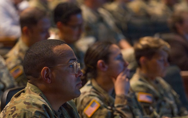 A large group of U.S. Army soldiers in OCP (Operational Camouflage Pattern) uniforms are seated in an auditorium-style venue, attentively watching a presentation or briefing. The foreground soldier is wearing glasses and holding a small notebook, suggesting active note-taking. The setting appears to be a formal military conference, training event, or briefing session.