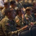 A large group of U.S. Army soldiers in OCP (Operational Camouflage Pattern) uniforms are seated in an auditorium-style venue, attentively watching a presentation or briefing. The foreground soldier is wearing glasses and holding a small notebook, suggesting active note-taking. The setting appears to be a formal military conference, training event, or briefing session.