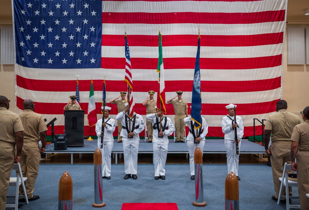 A formal U.S. Navy military ceremony is taking place indoors in front of a large American flag backdrop. A color guard in white dress uniforms presents flags including the U.S. flag, what appears to be the Italian flag, and a U.S. Navy command flag, while officers in khaki uniforms render salutes on stage. Decorative torpedo or missile models are positioned on the floor flanking a red carpet runner.