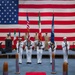 A formal U.S. Navy military ceremony is taking place indoors in front of a large American flag backdrop. A color guard in white dress uniforms presents flags including the U.S. flag, what appears to be the Italian flag, and a U.S. Navy command flag, while officers in khaki uniforms render salutes on stage. Decorative torpedo or missile models are positioned on the floor flanking a red carpet runner.