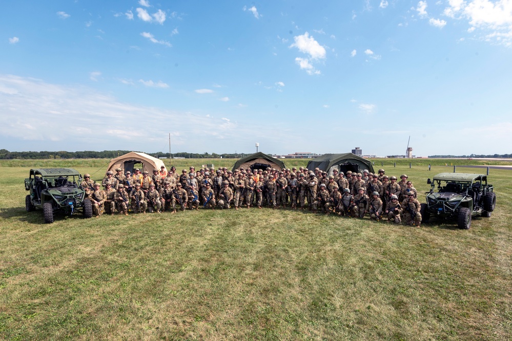 A large group of military personnel in camouflage uniforms poses for a formal group photograph on an open grass field, likely at or near an airfield. The group is flanked by two military utility terrain vehicles (UTVs) and backed by several military field tents. The setting appears to be a training exercise or field operation based on the equipment and infrastructure visible.