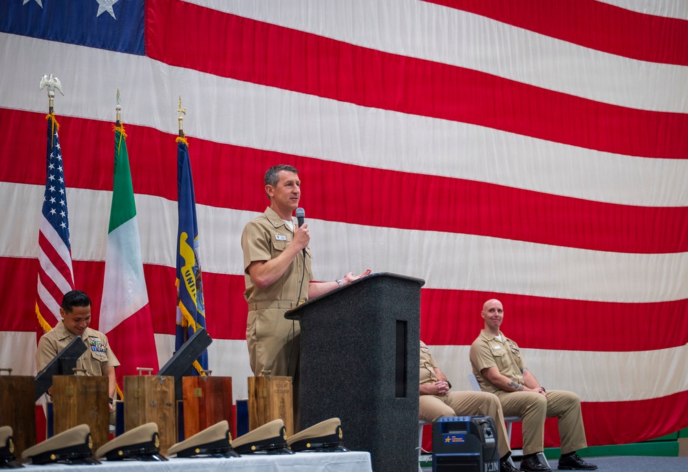 A formal U.S. Navy military ceremony is taking place on a stage in front of a large American flag backdrop. A naval officer in tan/khaki service uniform is speaking at a podium with a microphone, while two other personnel are seated or standing nearby. Flags including the U.S. flag and what appears to be the Italian flag are displayed on the stage.