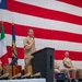 A formal U.S. Navy military ceremony is taking place on a stage in front of a large American flag backdrop. A naval officer in tan/khaki service uniform is speaking at a podium with a microphone, while two other personnel are seated or standing nearby. Flags including the U.S. flag and what appears to be the Italian flag are displayed on the stage.