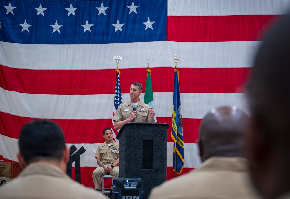 A U.S. Navy officer in tan/khaki service uniform is speaking at a podium during a formal military ceremony. A large American flag serves as a backdrop, flanked by what appears to be a Mexican flag and a U.S. Navy command flag. An audience of uniformed personnel is visible in the foreground.