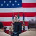 A U.S. Navy officer in tan/khaki service uniform is speaking at a podium during a formal military ceremony. A large American flag serves as a backdrop, flanked by what appears to be a Mexican flag and a U.S. Navy command flag. An audience of uniformed personnel is visible in the foreground.