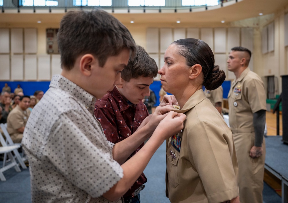 A military promotion or advancement ceremony is taking place in what appears to be a gymnasium or large hall. Two young boys, likely family members, are pinning a new insignia or rank device onto the collar of a female U.S. Navy sailor in tan service uniform. Additional uniformed personnel and seated audience members are visible in the background.