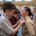 A military promotion or advancement ceremony is taking place in what appears to be a gymnasium or large hall. Two young boys, likely family members, are pinning a new insignia or rank device onto the collar of a female U.S. Navy sailor in tan service uniform. Additional uniformed personnel and seated audience members are visible in the background.