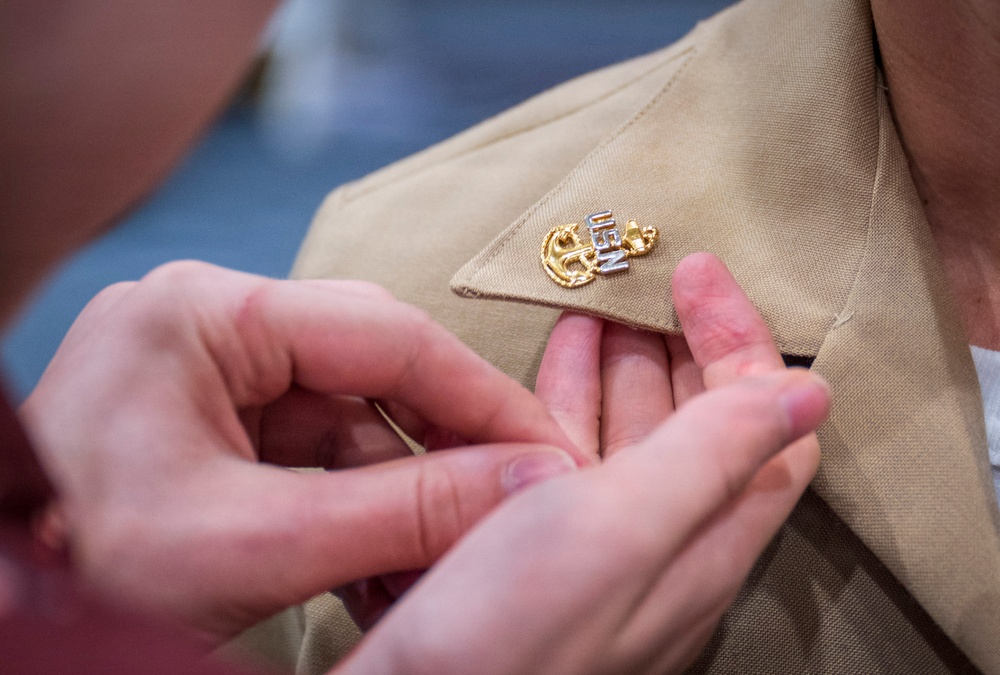 A close-up photograph shows hands pinning or adjusting a military insignia onto a tan/khaki uniform collar. The badge appears to be a U.S. Navy Chief Petty Officer collar device, featuring a gold anchor with silver 'USN' lettering. This appears to be a pinning ceremony, a traditional military promotion or advancement ritual.