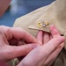 A close-up photograph shows hands pinning or adjusting a military insignia onto a tan/khaki uniform collar. The badge appears to be a U.S. Navy Chief Petty Officer collar device, featuring a gold anchor with silver 'USN' lettering. This appears to be a pinning ceremony, a traditional military promotion or advancement ritual.