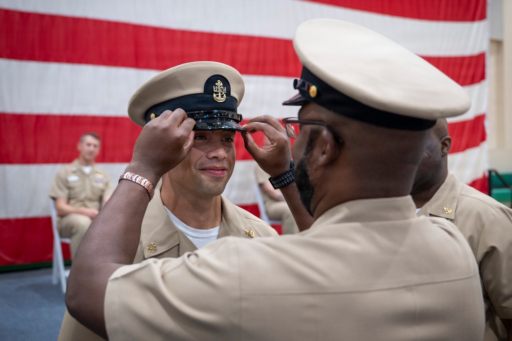 A U.S. Navy Chief Petty Officer pinning ceremony is taking place, where a senior enlisted sailor is placing a combination cover (cap) on a newly promoted Chief Petty Officer. The ceremony is conducted in front of a large American flag backdrop, consistent with a formal military advancement ritual. Multiple sailors in tan service uniforms are present as witnesses.