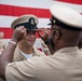 A U.S. Navy Chief Petty Officer pinning ceremony is taking place, where a senior enlisted sailor is placing a combination cover (cap) on a newly promoted Chief Petty Officer. The ceremony is conducted in front of a large American flag backdrop, consistent with a formal military advancement ritual. Multiple sailors in tan service uniforms are present as witnesses.
