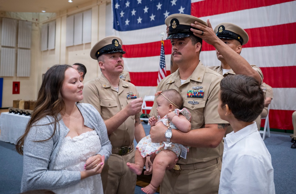 A U.S. Navy promotion ceremony is taking place in front of a large American flag. A sailor holding an infant is having his chief petty officer cover (hat) placed on his head, while his family — including a woman and a young boy — stand nearby. The scene captures a formal military milestone shared with loved ones.