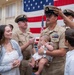 A U.S. Navy promotion ceremony is taking place in front of a large American flag. A sailor holding an infant is having his chief petty officer cover (hat) placed on his head, while his family — including a woman and a young boy — stand nearby. The scene captures a formal military milestone shared with loved ones.